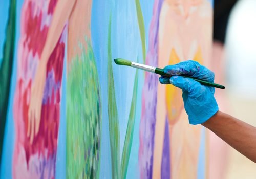 Artist's hand with paintbrush painting colorful picture on canvas at outdoor art festival. Woman painter in blue gloves draws surreal picture with paintbrush, beautiful art performance