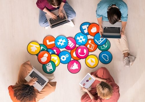 Top view of four young friendly people in casualwear sitting by multi-color papers with icons on the floor and using mobile gadgets