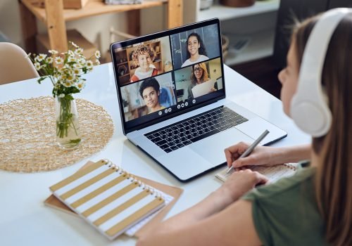 Over shoulder view of student girl in headphones sitting at table and participating in online conference in student group